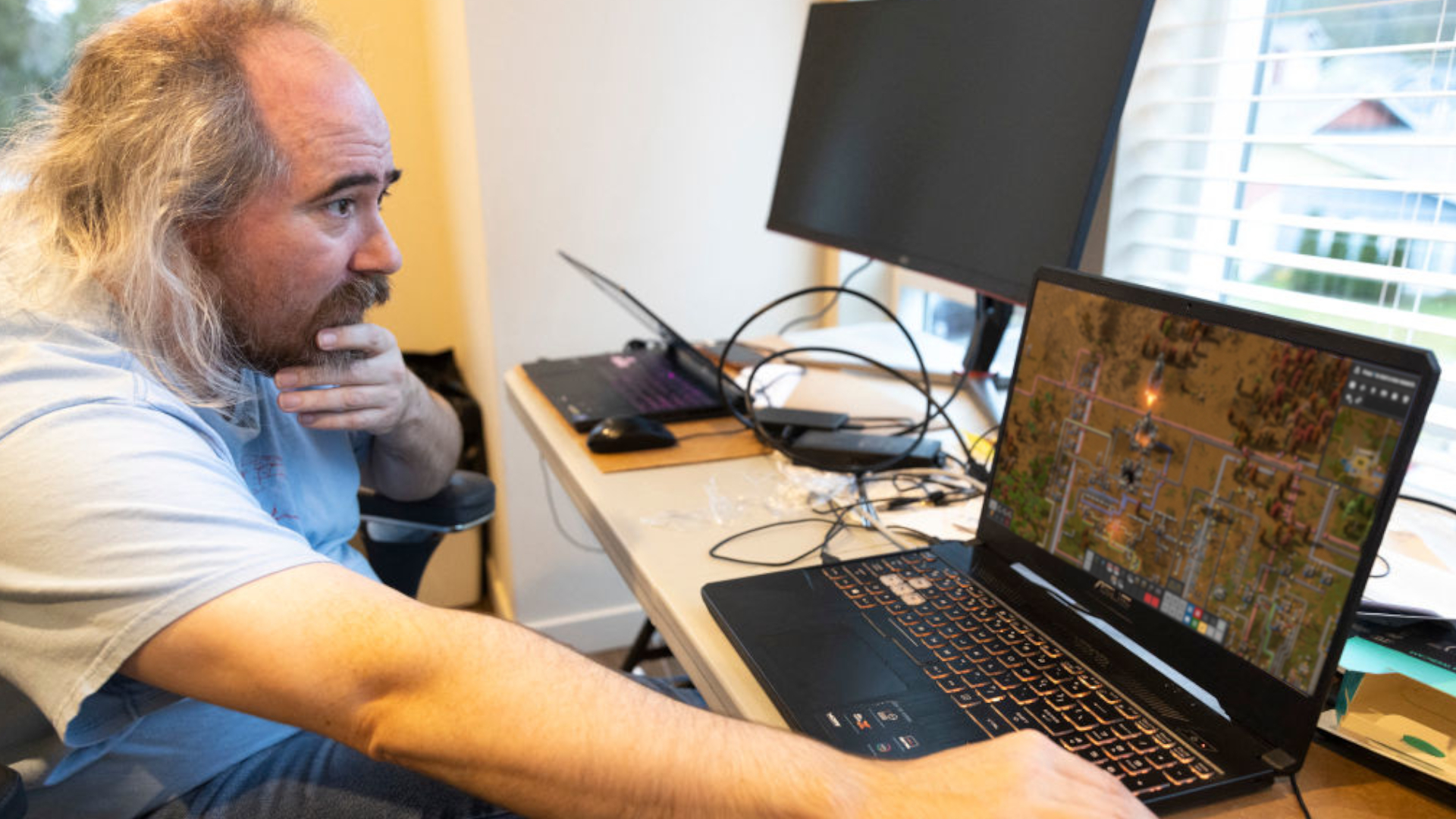 Tarn Adams sitting in front of a laptop, edited with a screen from Factorio