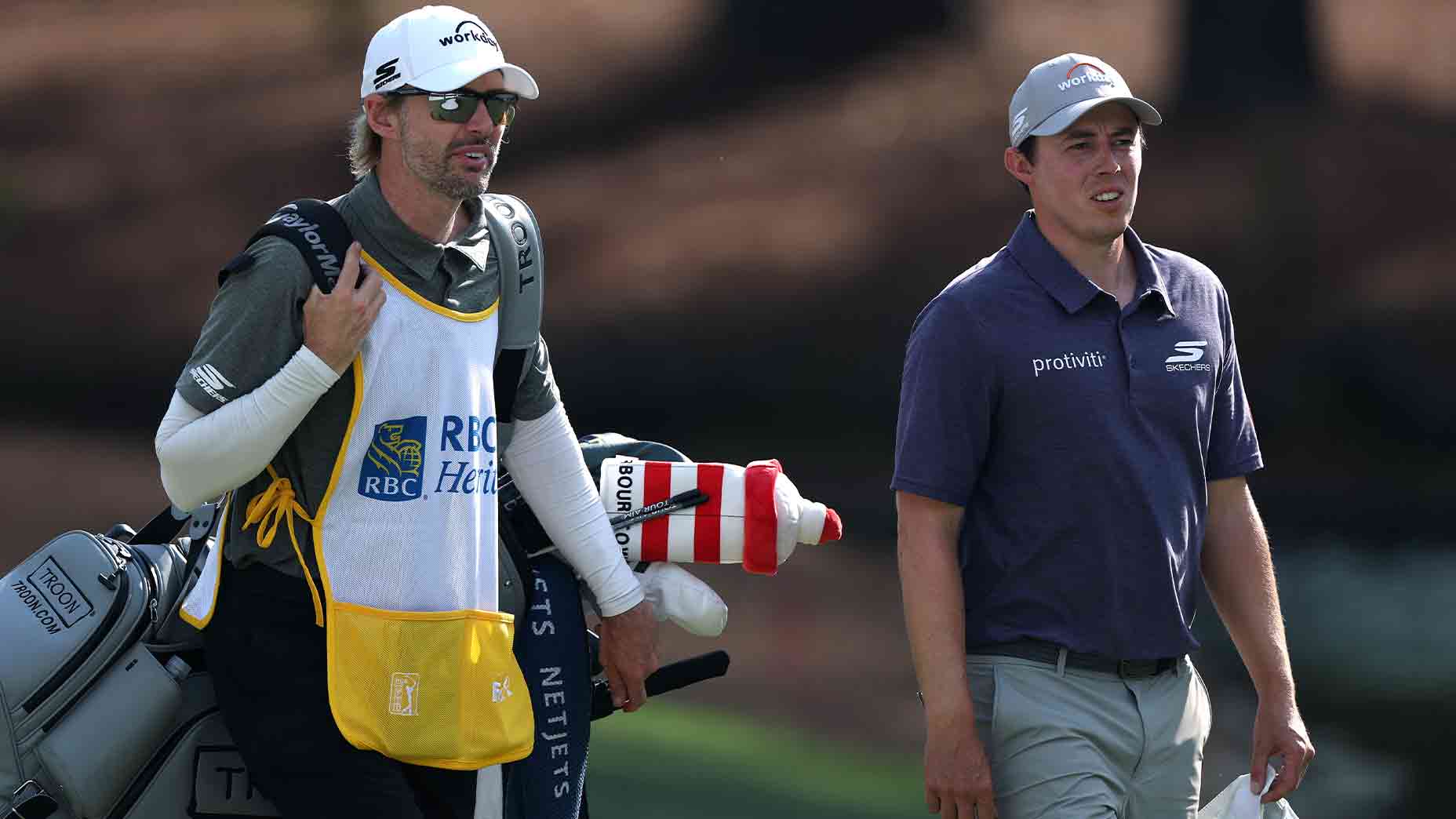Two men walk side by side on a golf course during the 2026 RBC Heritage. One, in sunglasses and a white caddie bib, carries golf clubs; the other, in a purple polo and gray cap, is the golfer. Sunlight highlights their faces as they walk.