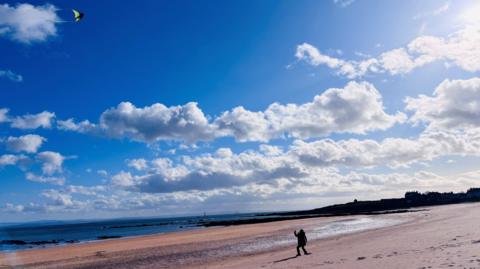 A wide sandy beach under a bright blue sky with scattered clouds. A person walks along the shore while a colourful kite flies high above.