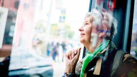 An older woman smiles as she looks out of a bus window. She has short grey hair and is wearing a green scarf.