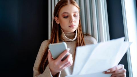 Young woman with red hair, wearing beige jumper holds smartphone in one hand and household bills in the other