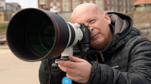 Owen Humphreys is taking a picture with his long lens camera. He has one eye behind the device, with the other looking at the beach scene ahead. Humphreys is bald and is wearing a single black earring and a black puffer jacket.