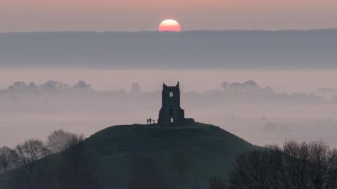 A silhouetted image taken from a distance, showing a group of people enjoying a misty sunrise over the monument at Burrow Mump in Somerset.