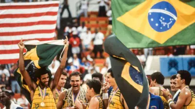 A equipe do Brasil comemora a vitória na partida de basquete que lhe rendeu a medalha de ouro contra os Estados Unidos durante os Jogos Pan-Americanos de 1987, realizados em agosto de 1987 na Market Square Arena em Indianápolis, Indiana; Oscar Schmidt aparece visível com a rede em volta do pescoço.