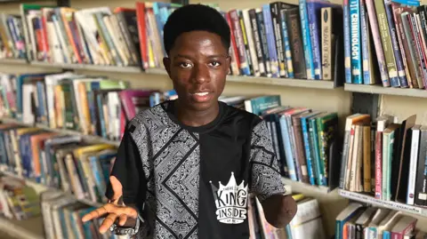 Ovey Friday stands in front of book shelves in a library in Nigeria. He is wearing black and white T-shirt and a large watch. One of his hands has fingers missing. The other arm shows a scar where his hand was amputated