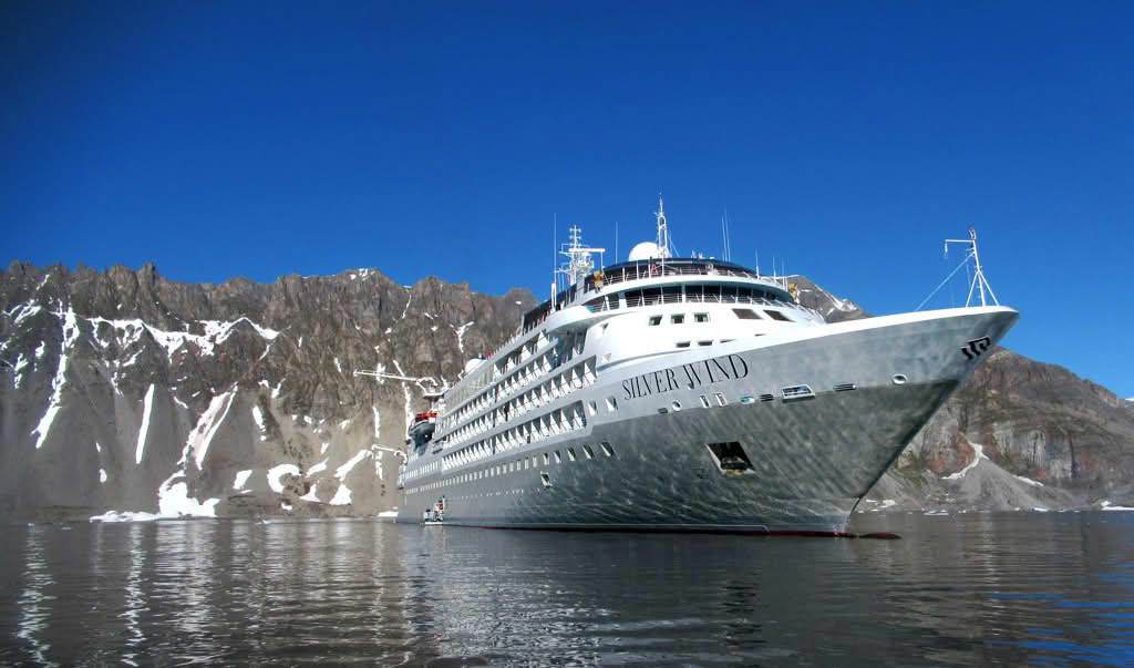 Foto av et stort cruiseskip som ligger til ro på stille vann. Baugen (foran) på skipet er nærmest kamera. Bak skipet er det bratte fjell med steinete skråninger som noen steder har flekker og striper av snø.